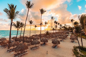 A beach with rows of sun loungers and thatched umbrellas, palm trees, and a clear view of the ocean.