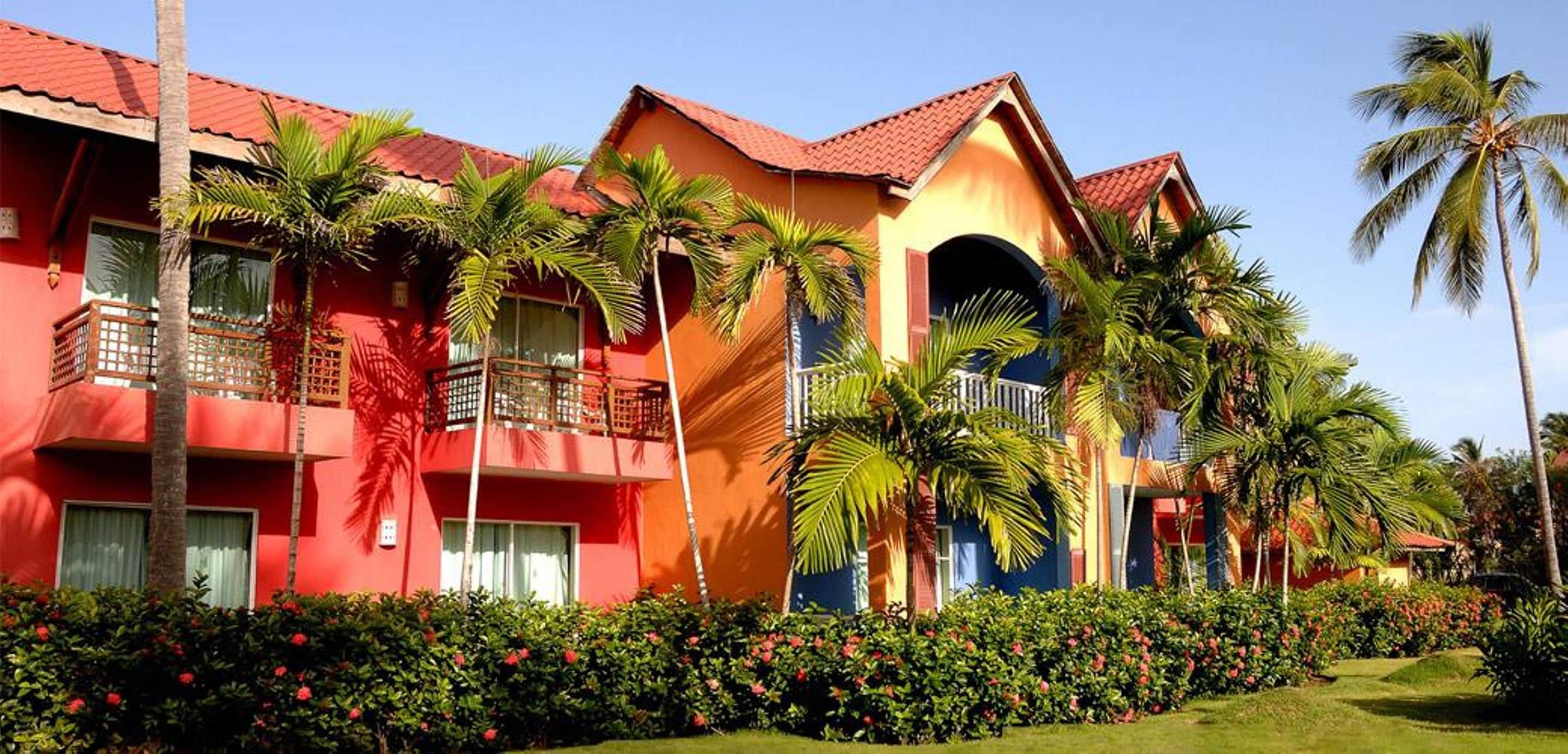 Un hotel con paredes exteriores rojas y amarillas, varios balcones y abundante vegetación, incluyendo palmeras y plantas con flores.