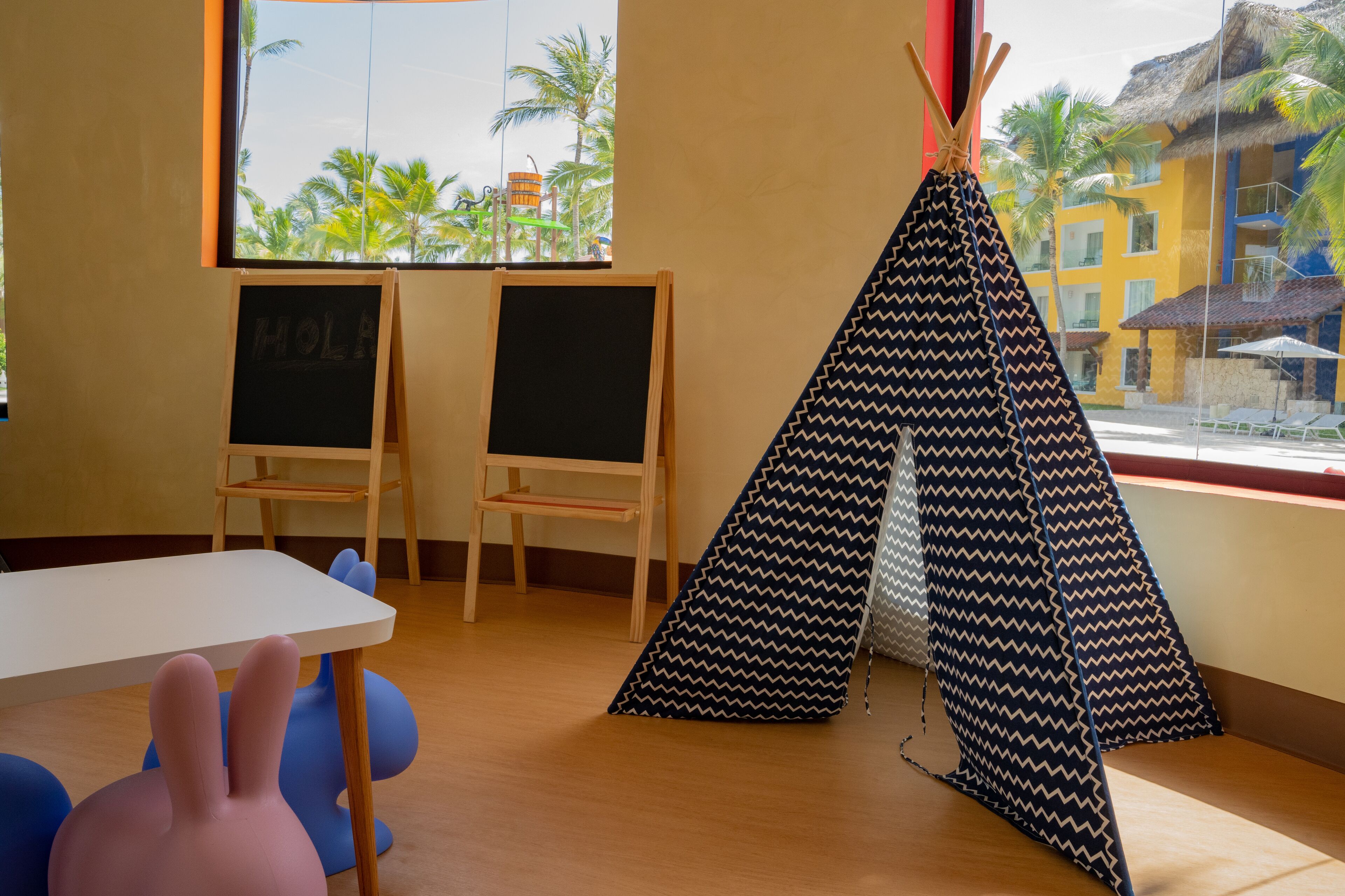 A room with a white table, two chalkboards, a blue and black patterned teepee, and a view of palm trees and buildings outside.