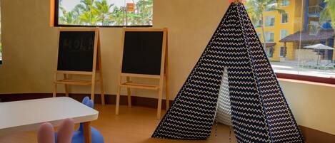 A room with a white table, two chalkboards, a blue and black patterned teepee, and a view of palm trees and buildings outside.