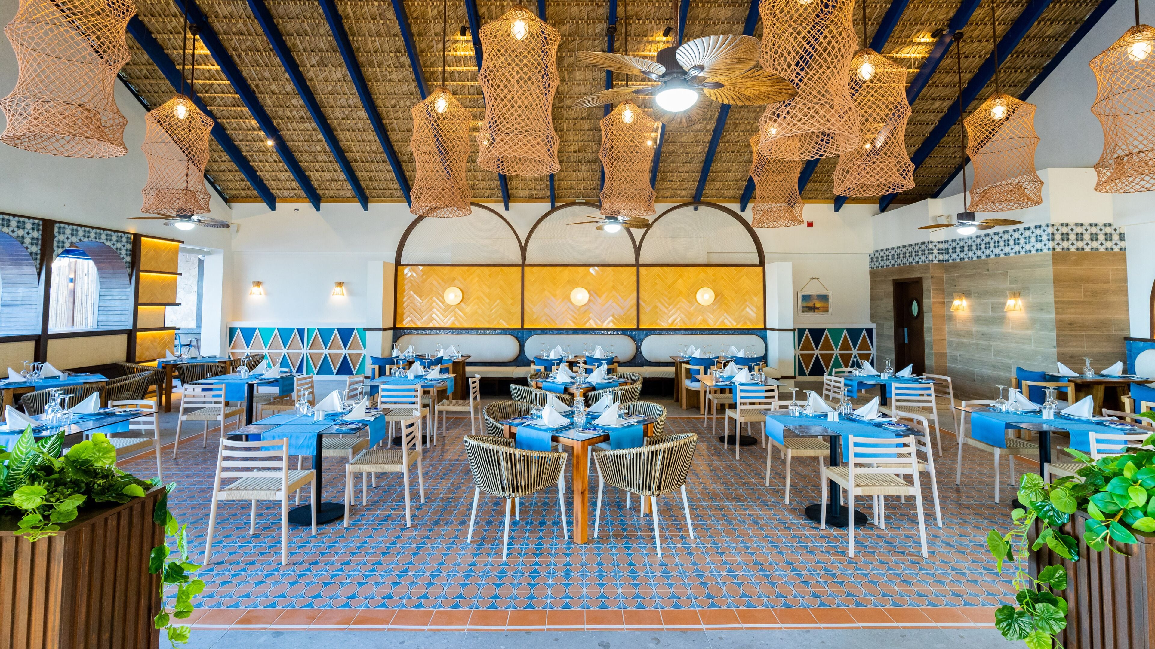 Ein geräumiges Restaurant mit blauen Tischdecken, Korbstühlen und einem gemusterten Boden.
