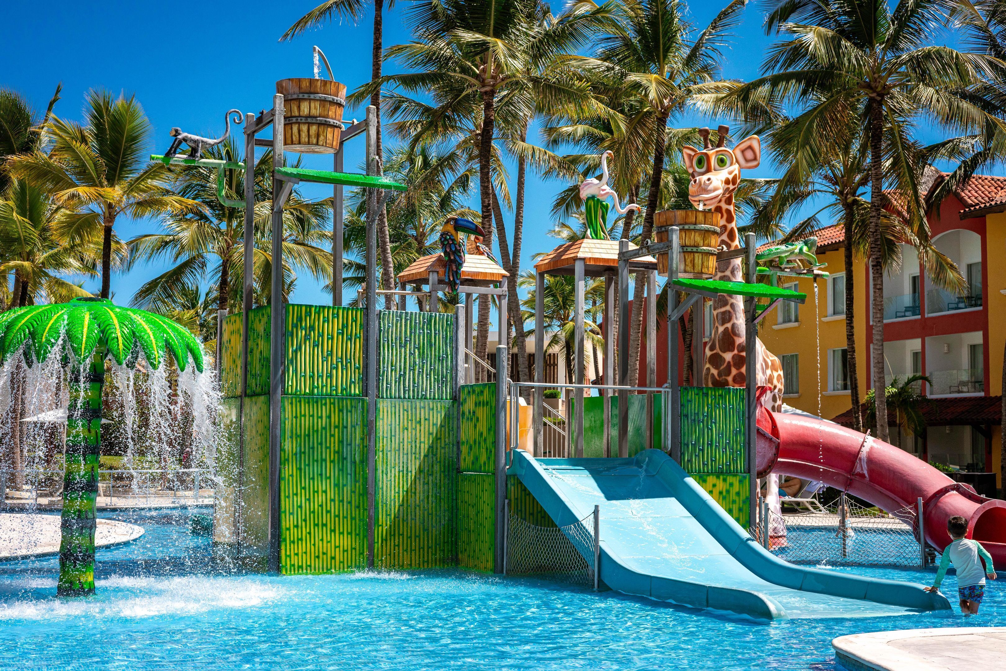 Een waterpark met glijbanen en een zwembad, omringd door palmbomen en een gebouw met een rood dak.