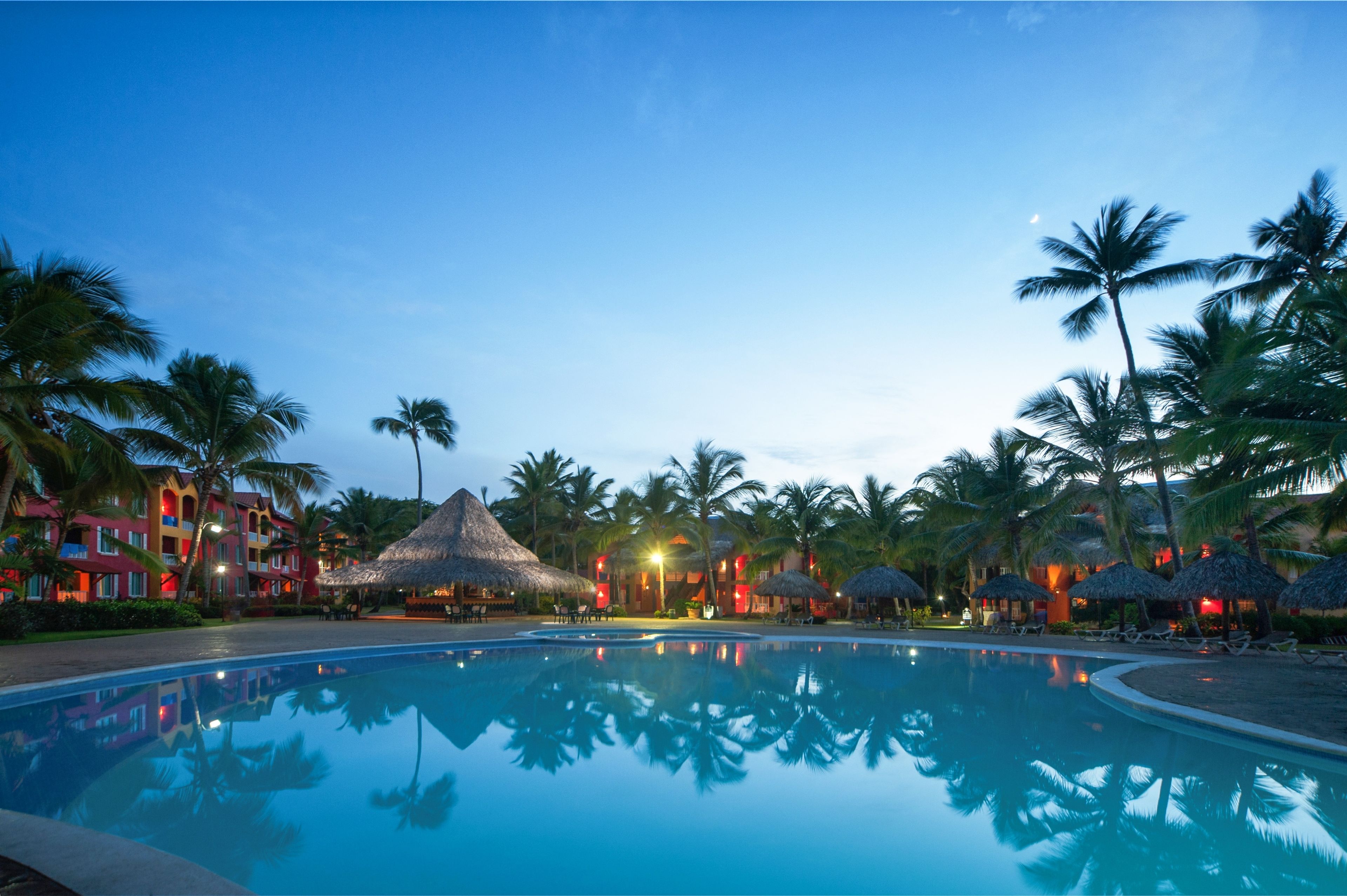 Una piscina de resort con palmeras y estructuras de techo de paja.