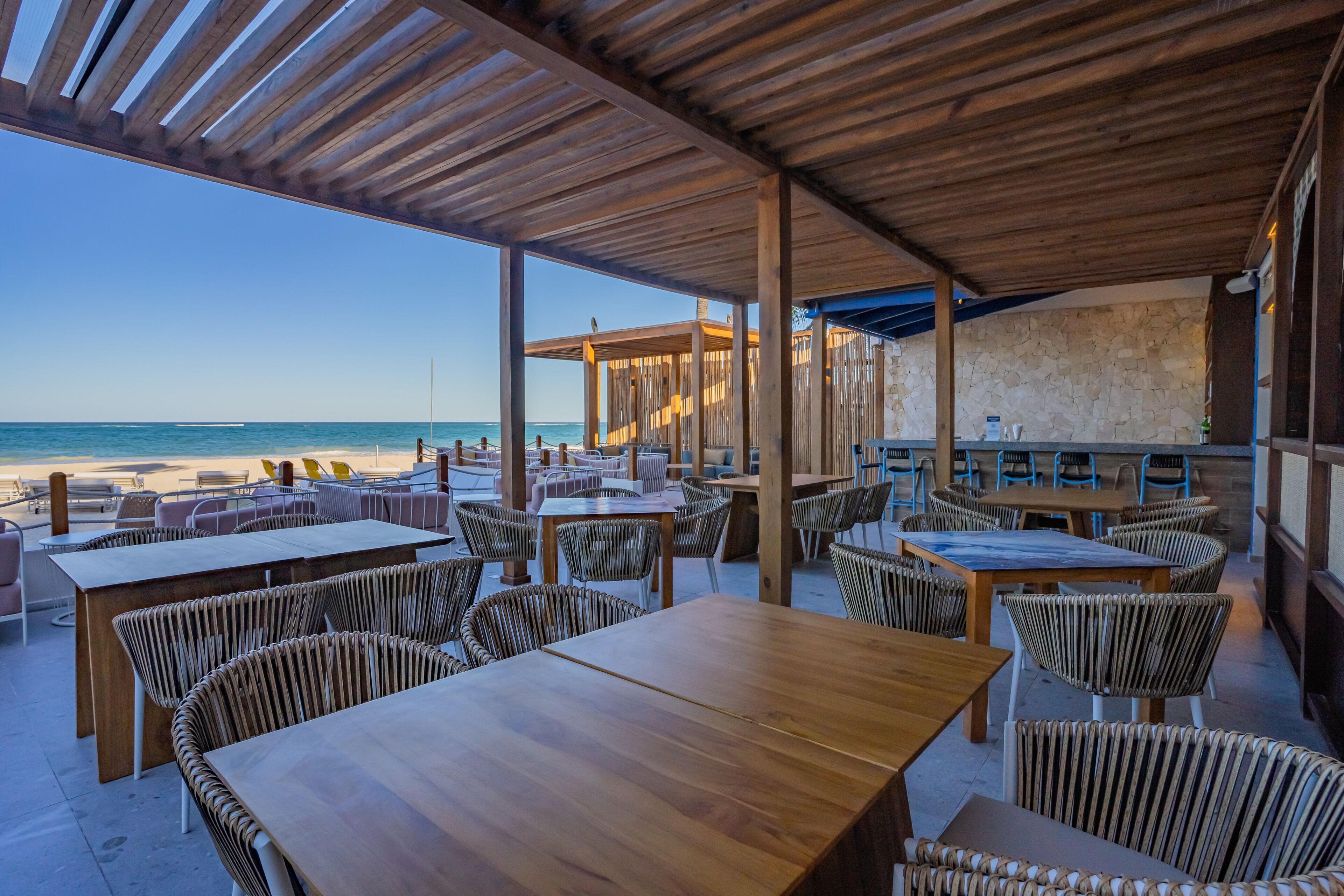 Ein Sitzbereich im Freien mit Holztischen und Korbstühlen mit Blick auf einen Strand.