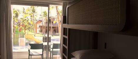 A room with a bed, a desk, and a chair. There is a sliding glass door leading to an outdoor area with a pool and palm trees.