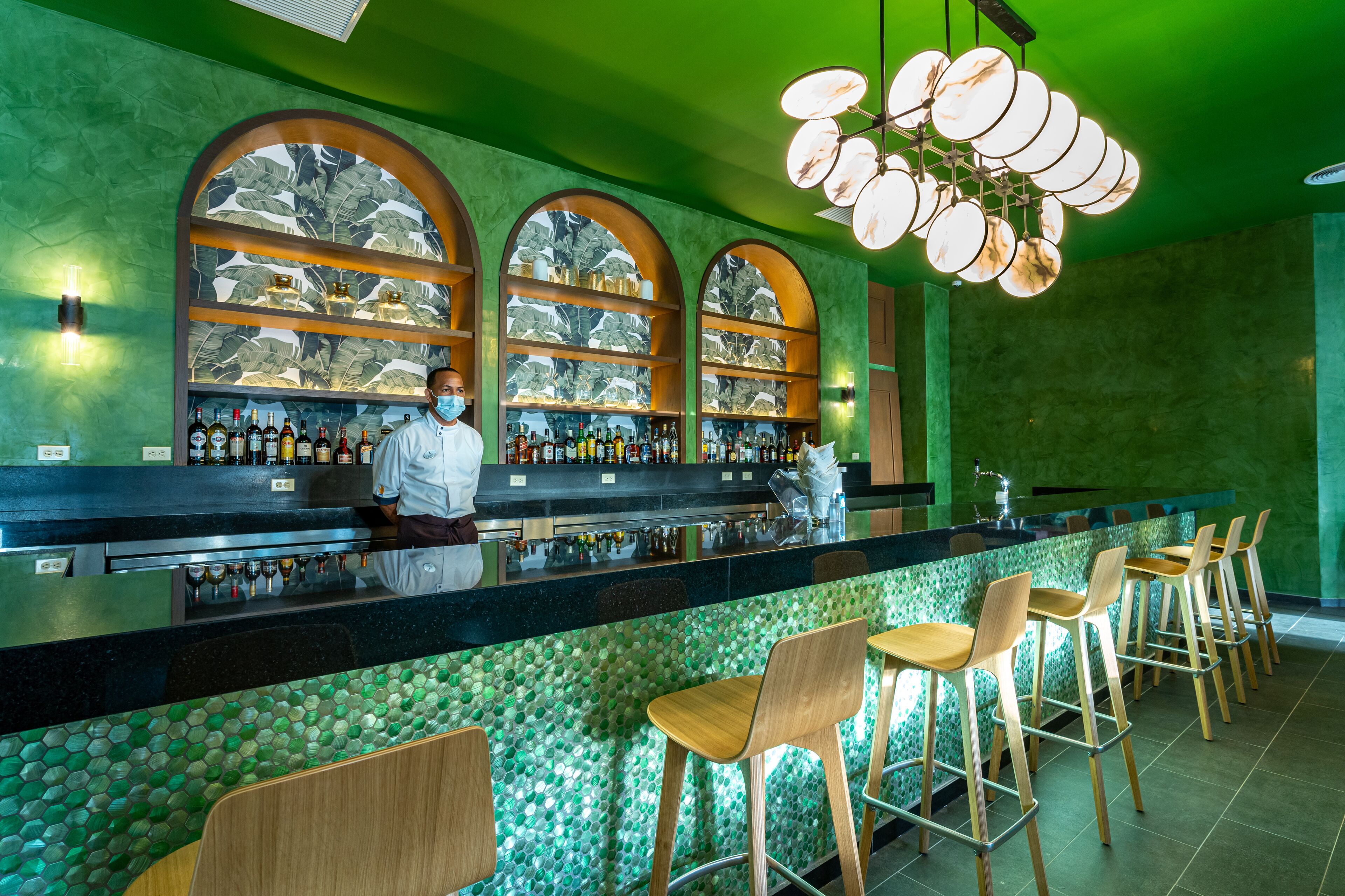 A bar area with a green-tiled counter, wooden bar stools, and a bartender wearing a mask.