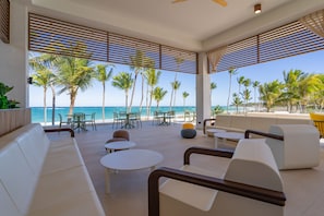 A modern outdoor lounge with white furniture, palm trees, and a view of the beach. - Tropical Deluxe Princess - All Inclusive (Punta Cana)