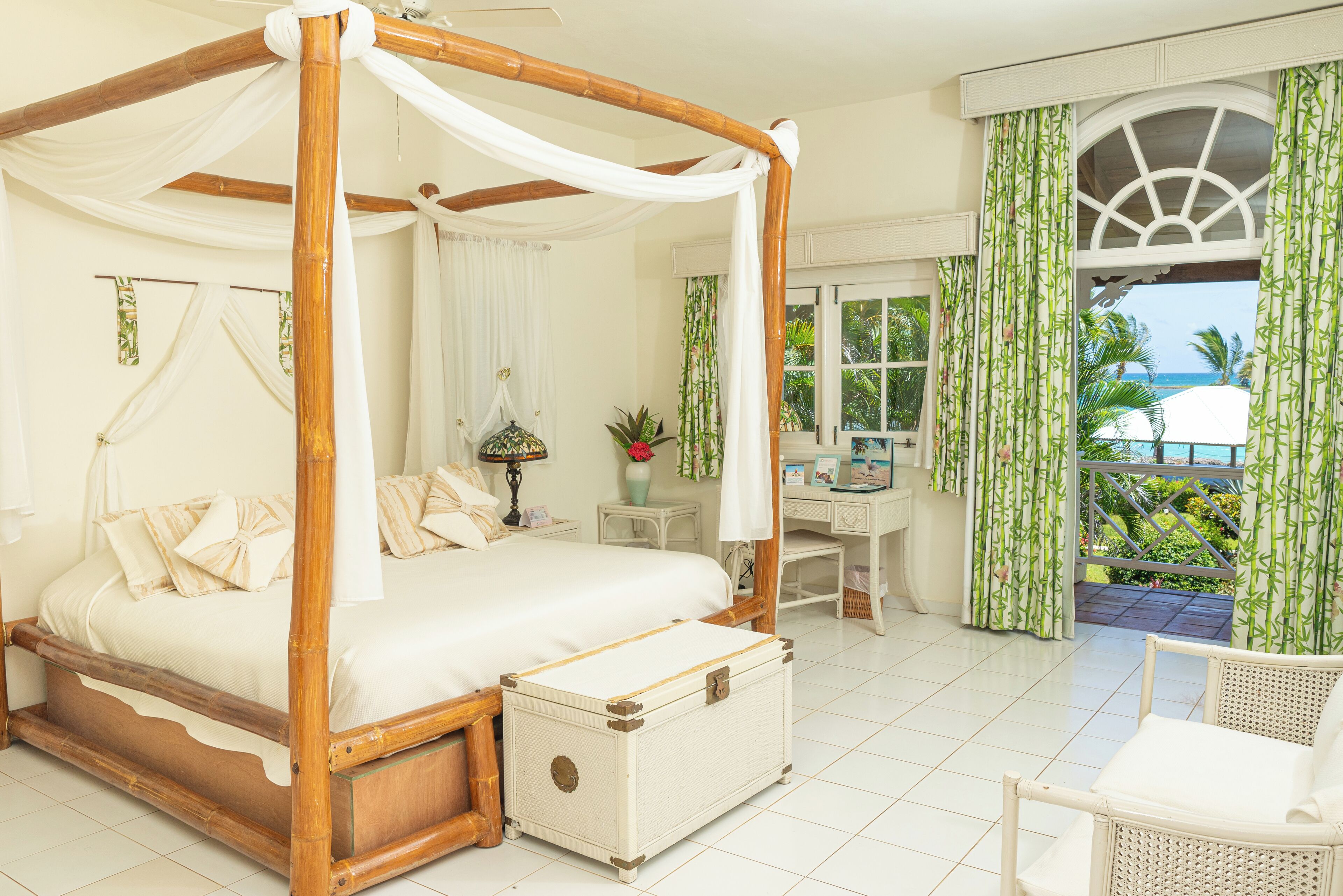 A four-poster bed with a canopy, a bedside table with a lamp, a small plant, and a view of the outdoors through a window with green curtains.