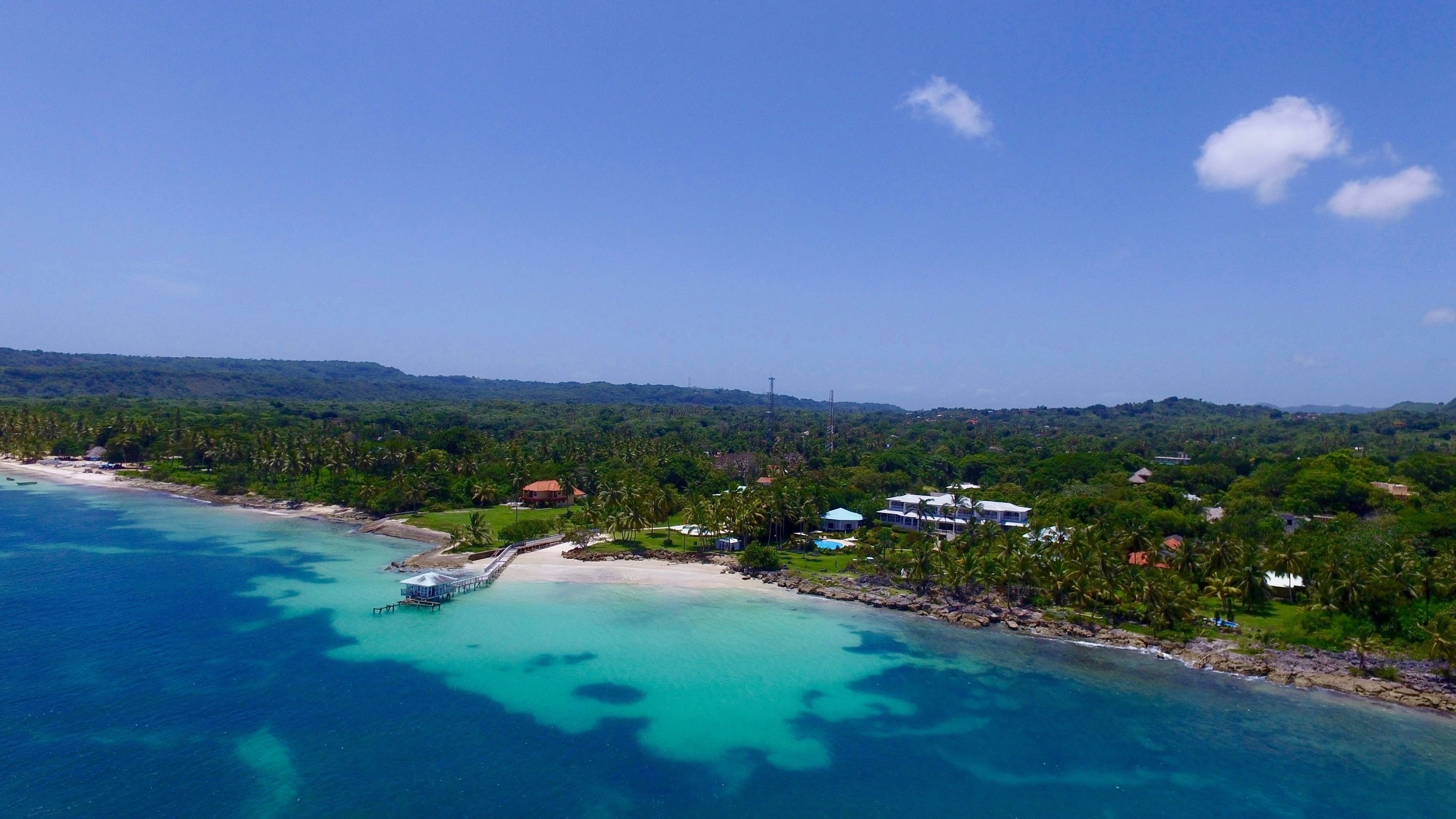 An aerial view of a coastal area with clear turquoise waters, a sandy beach, and a cluster of buildings surrounded by lush greenery.
