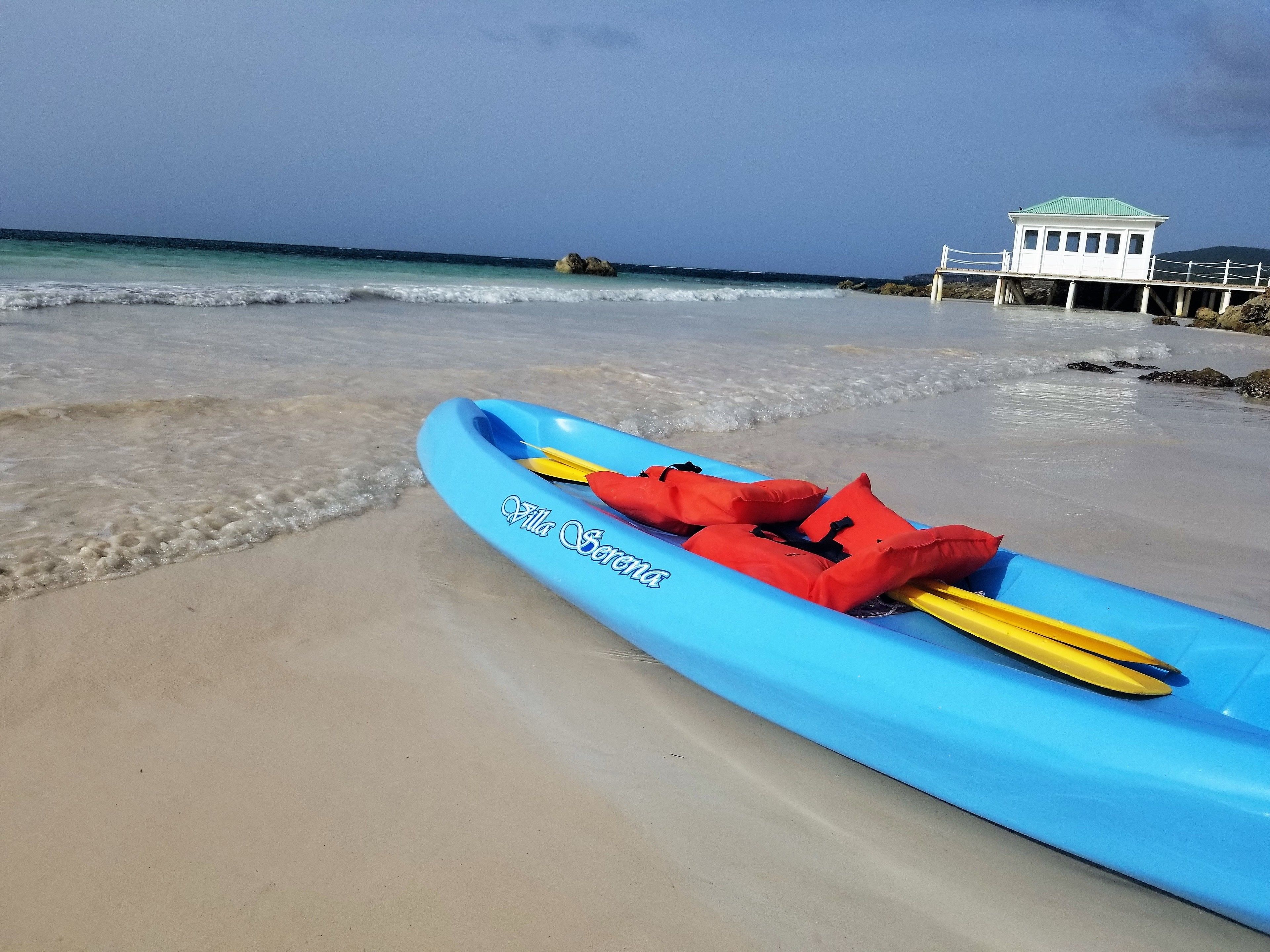 Un kayak blu con giubbotti di salvataggio rossi su una spiaggia sabbiosa, con un edificio sullo sfondo.