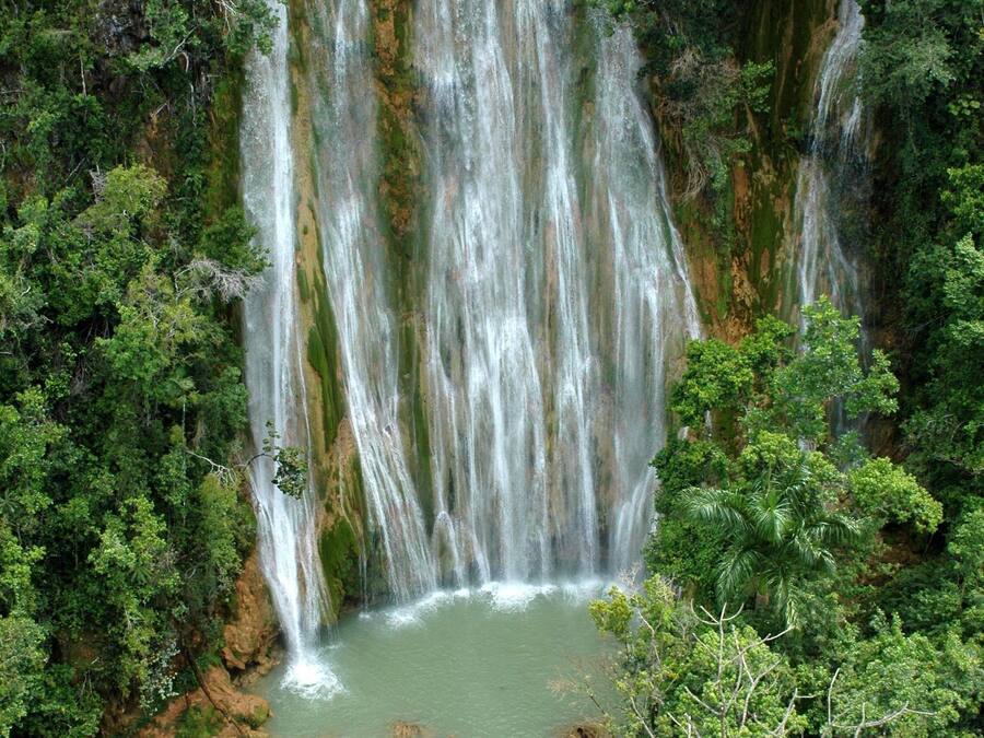 A waterfall surrounded by dense forest, with a river at the base and people swimming.