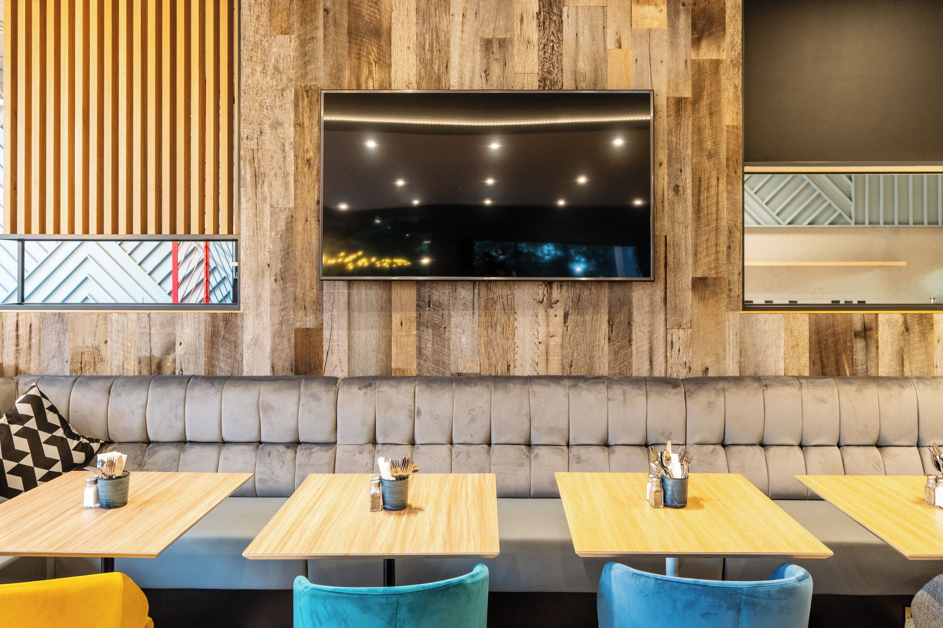 Een restaurantinterieur met houten paneelwanden, een flatscreen televisie boven een zachte bank en tafels met houten bladen en blauwe stoelen.