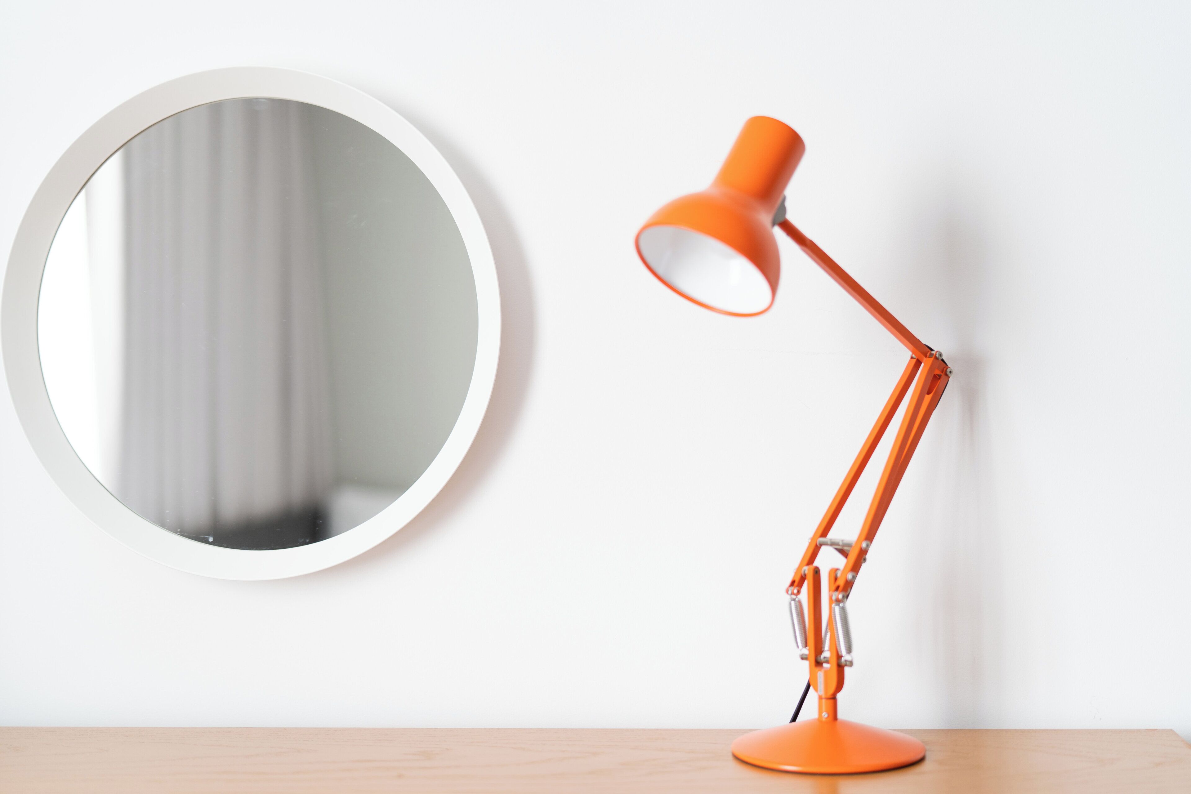 A round mirror and an orange desk lamp on a wooden surface against a white wall.