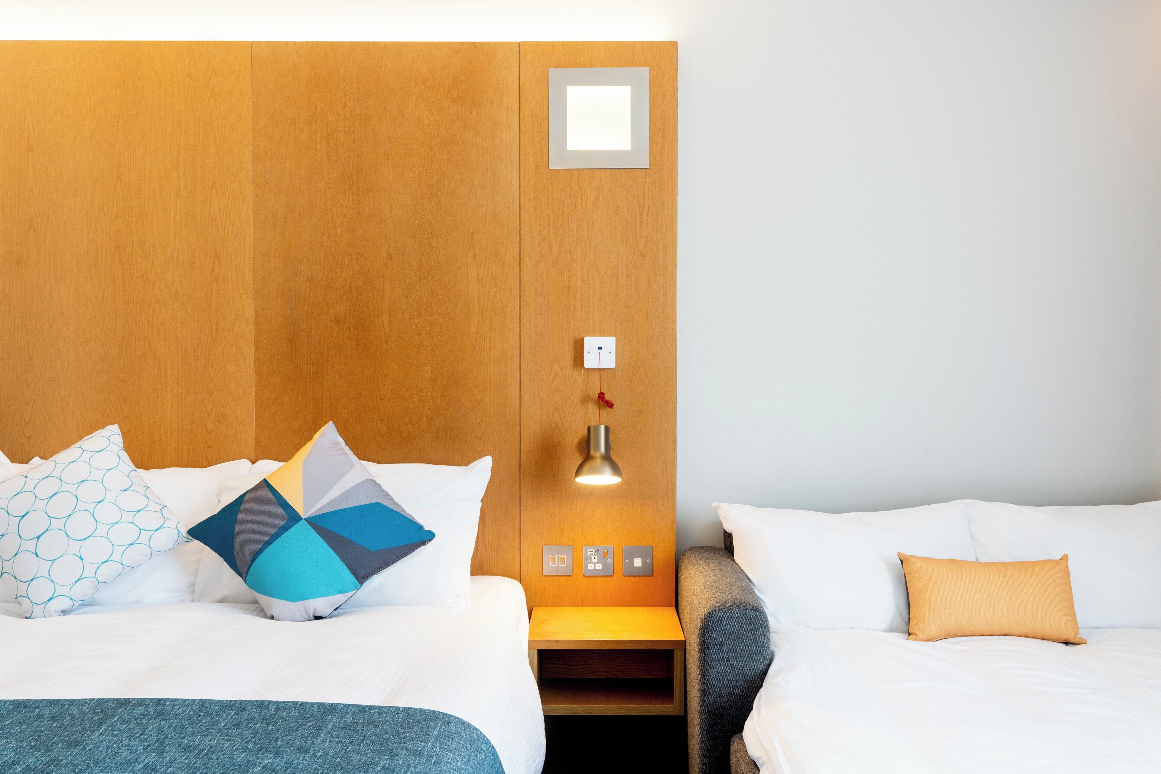 Habitació d'hotel amb dos llits, un reposacapses de fusta, una taula de nit amb una làmpada i una petita prestatgeria.
