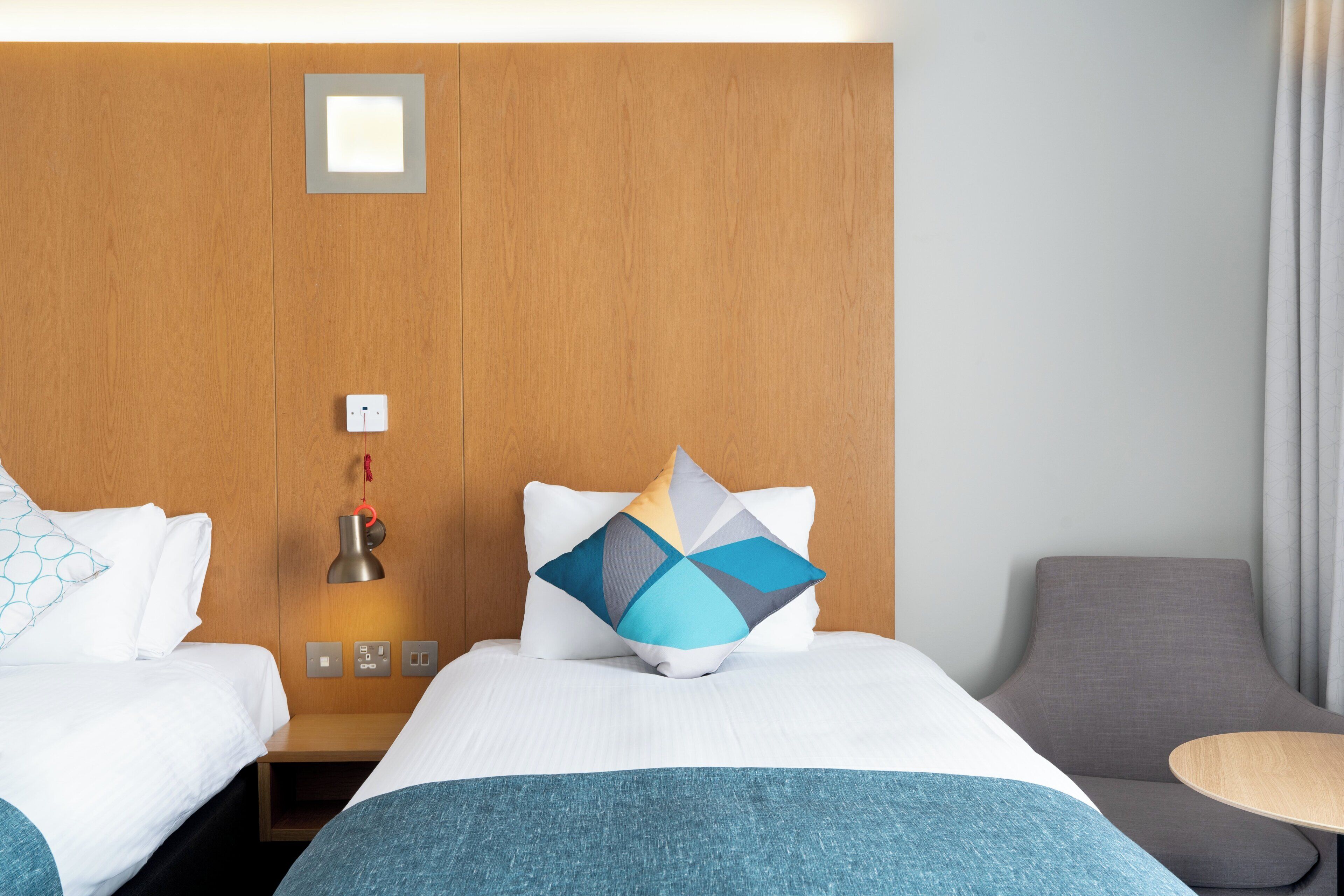 A hotel room with two beds, a wooden headboard, a bedside table with a lamp, and a grey armchair.