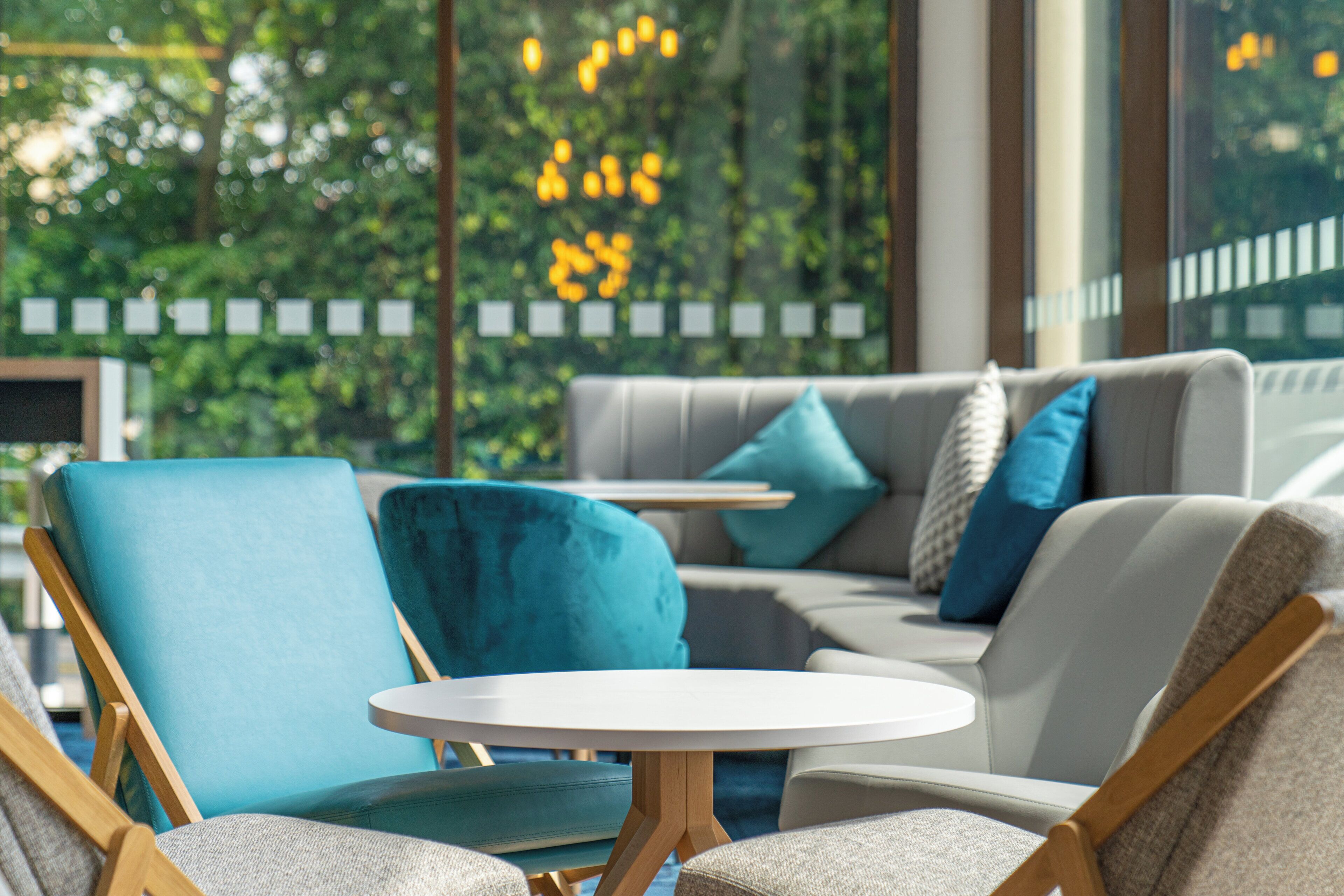 Een moderne zithoek met een bank en fauteuils, een klein rond tafeltje en uitzicht op groen via grote ramen.