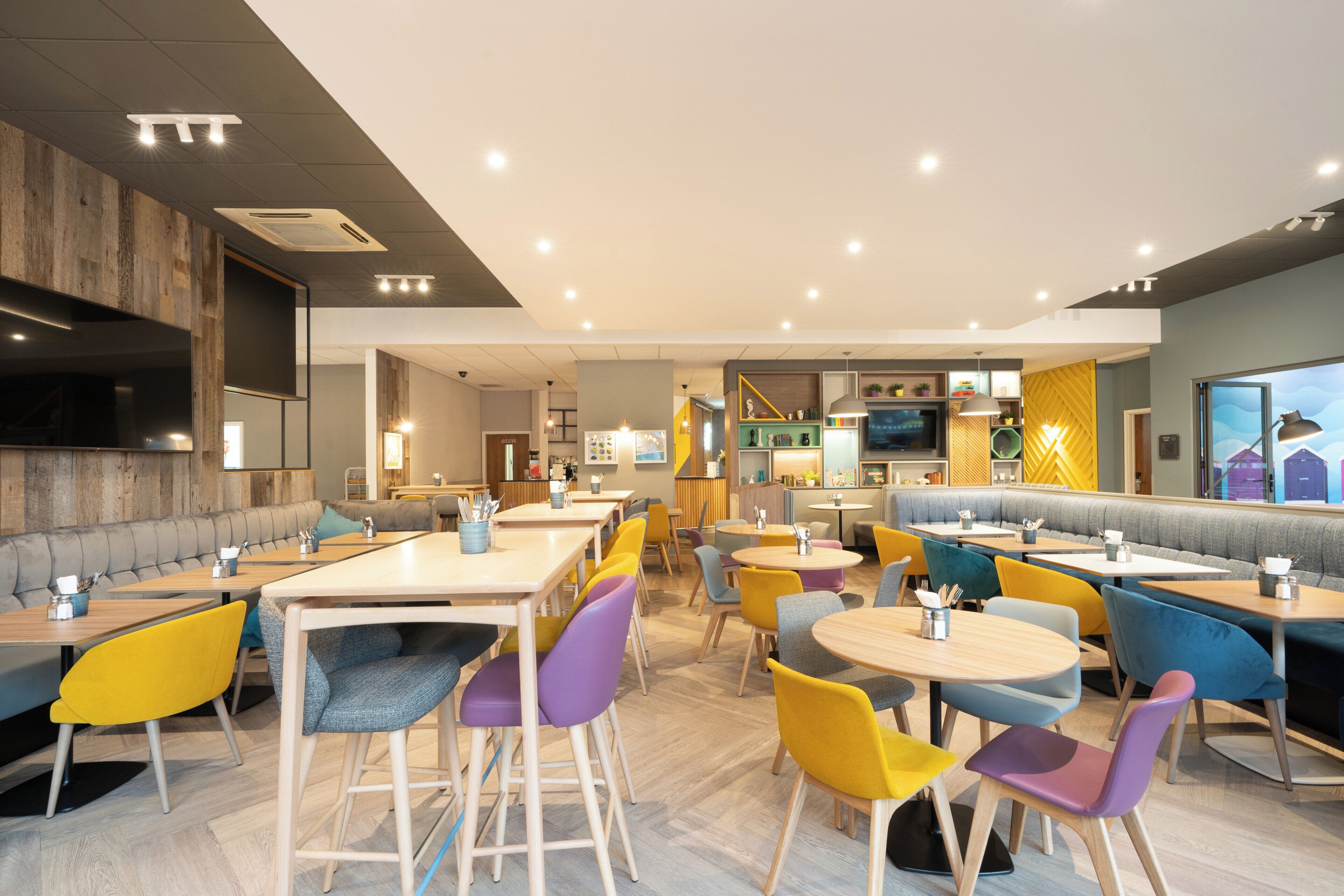 A modern restaurant interior with multiple seating options including booths and tables, a variety of colorful chairs, and a central dining area with a high ceiling and recessed lighting.