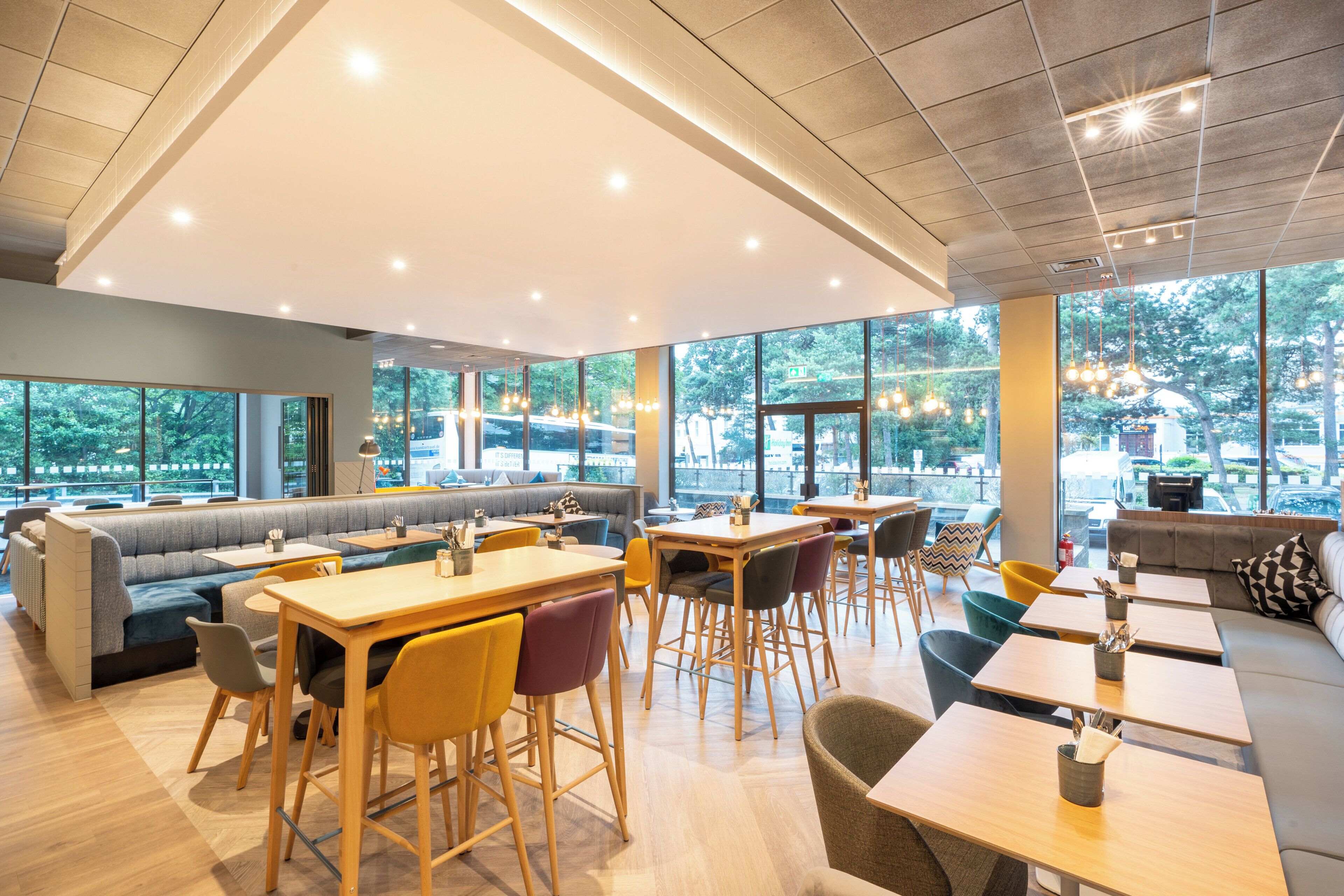 Un café moderne avec des espaces aménagés pour s’asseoir, de grandes fenêtres et une vue sur l’extérieur.