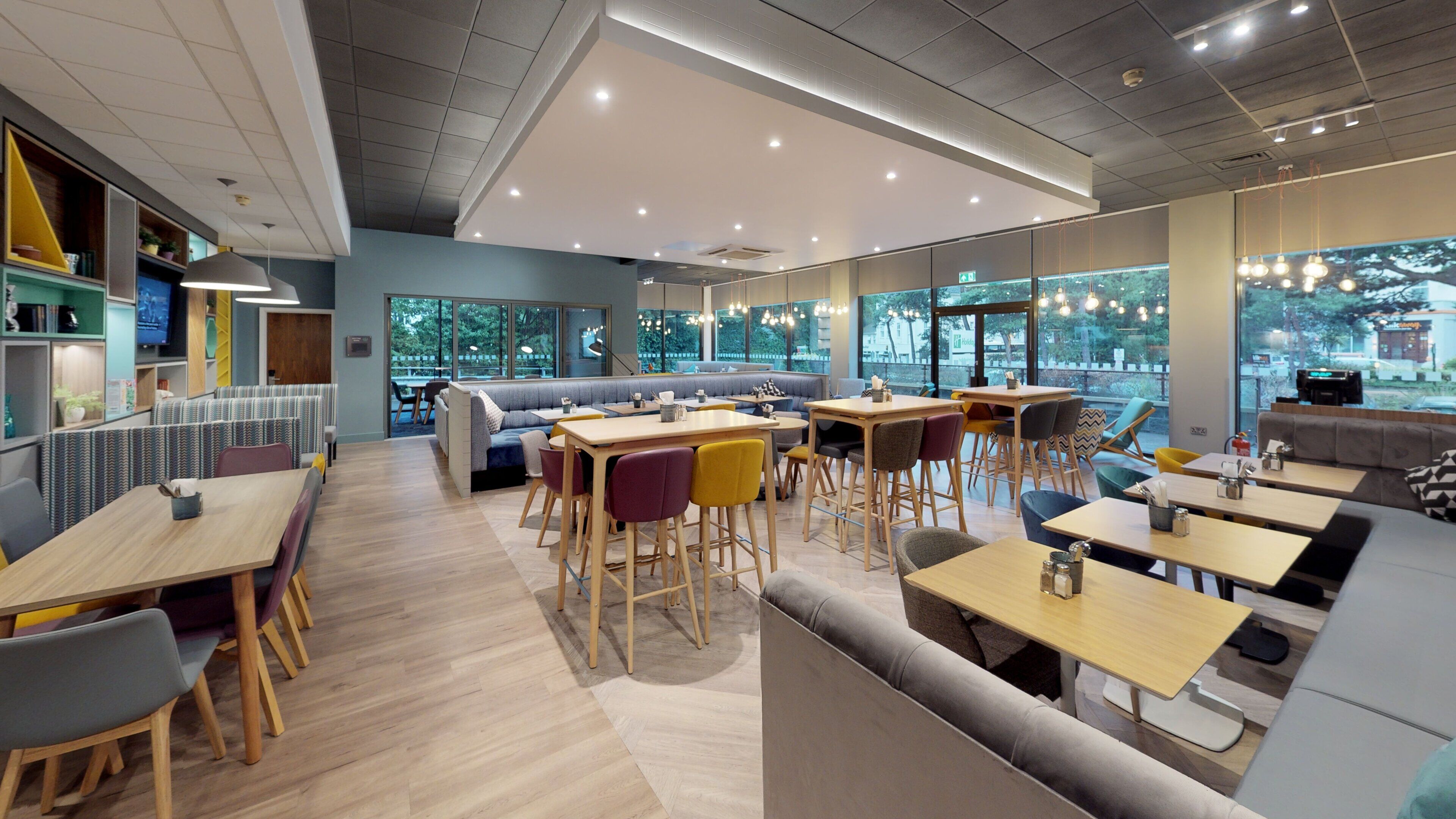Un café moderno con mesas y sillas de madera, un rincón con sofá y amplias ventanas que ofrecen vistas al exterior.