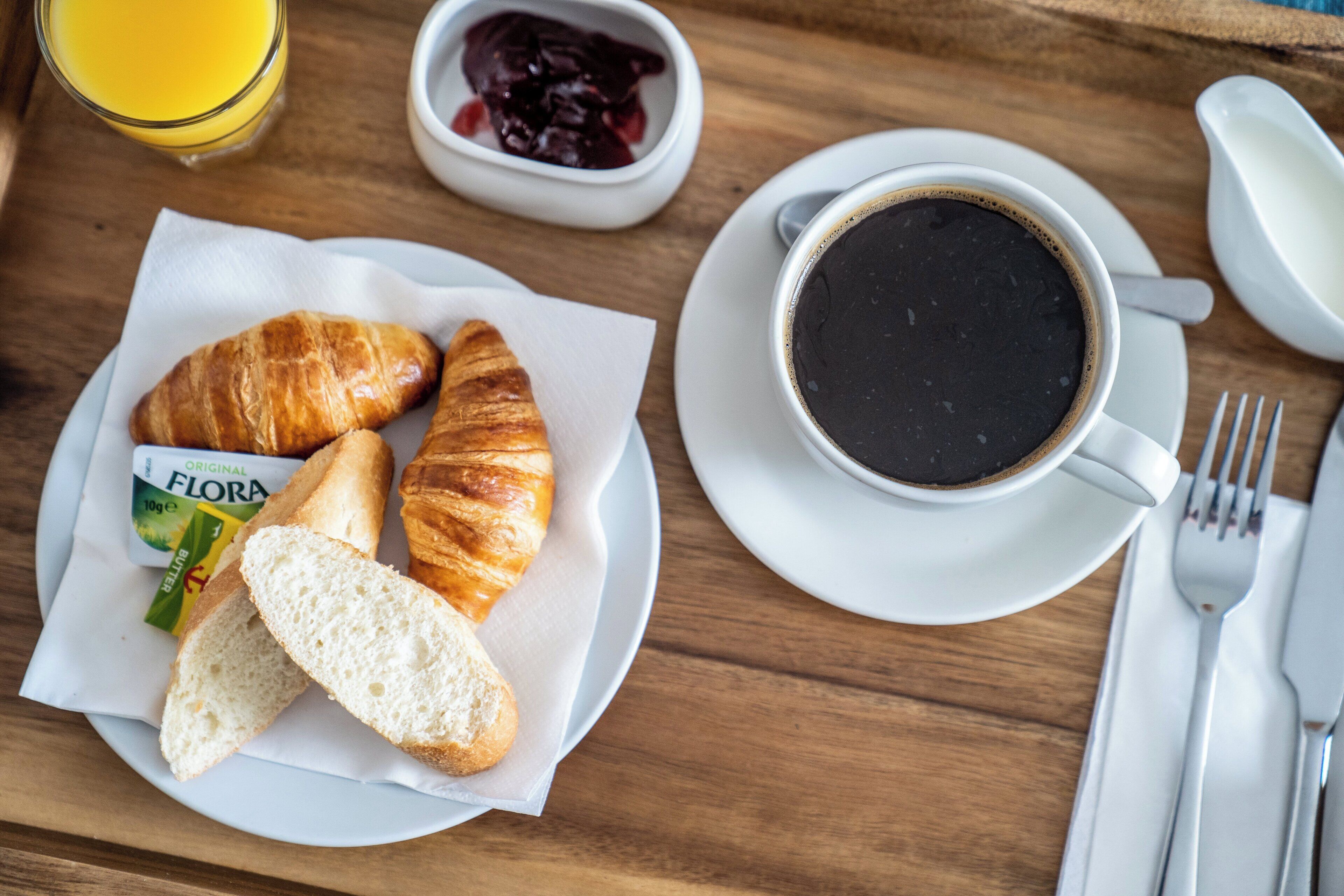 A breakfast setting with croissants, bread, a bowl of jam, a glass of orange juice, and a cup of coffee on a wooden table.