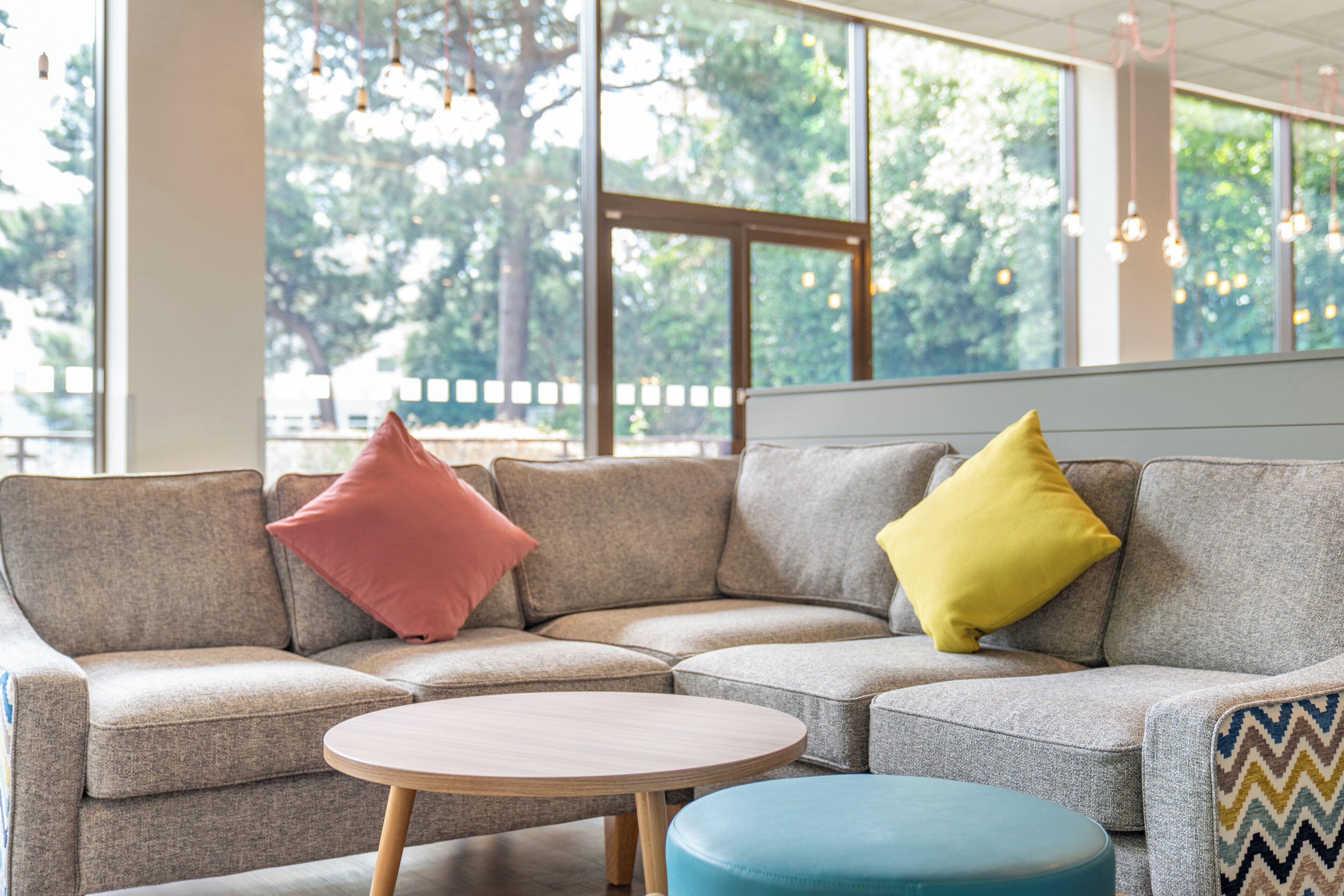 A sectional sofa with two pillows, a round wooden table, and a blue ottoman in a room with large windows and hanging lights.
