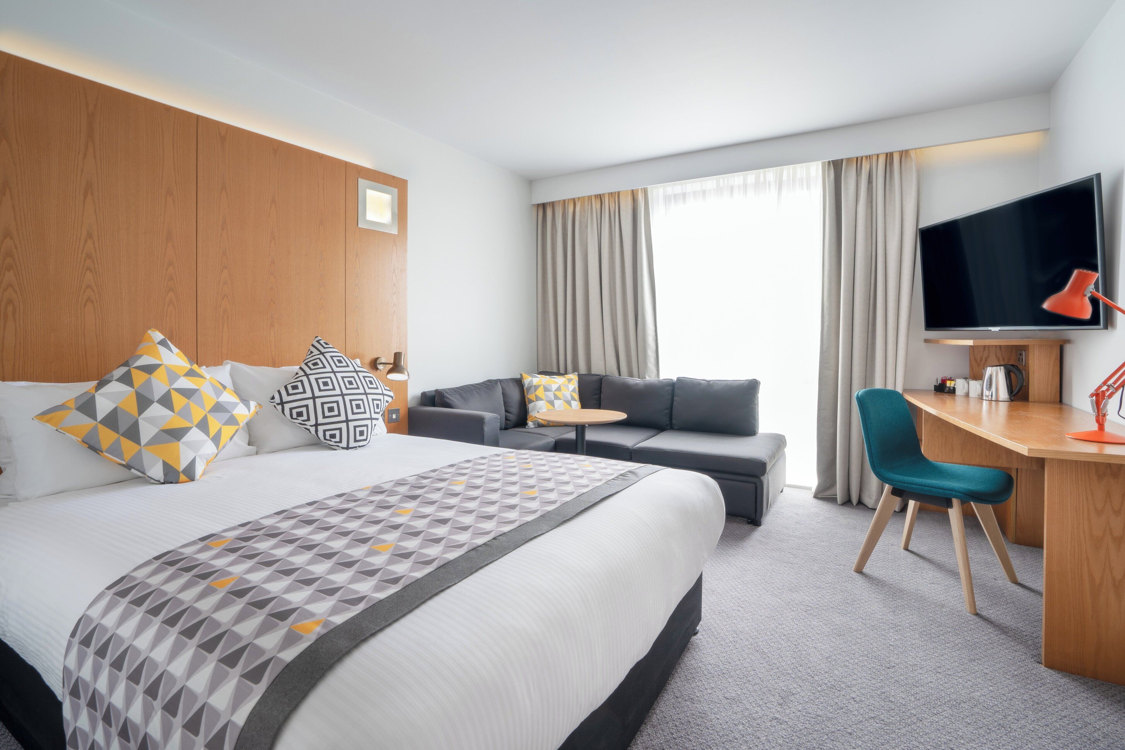 Ett hotellrum med en säng, en soffa, ett skrivbord, en stol, en tv och ett fönster med gardiner.