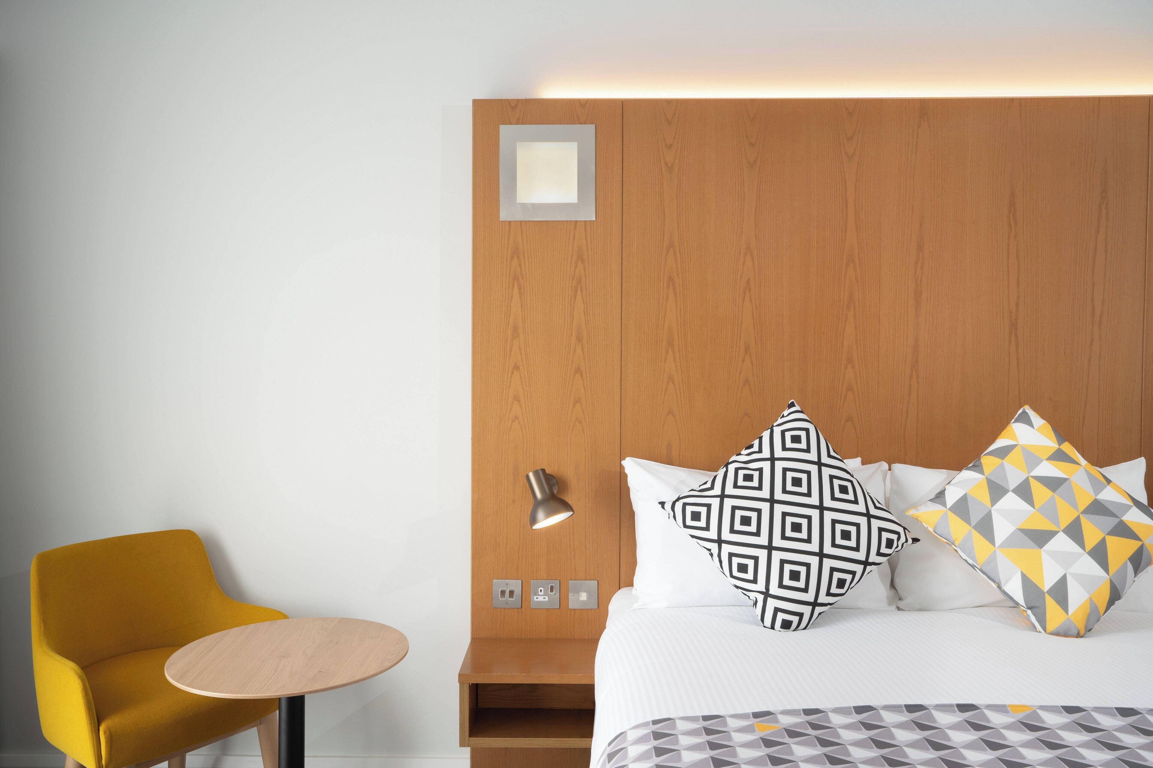 Une chambre d’hôtel avec une tête de lit en bois, un lit agrémenté d’oreillers à motifs, une petite table ronde et un fauteuil jaune.