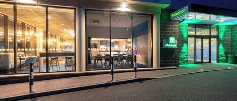 A hotel entrance with a reception desk, a glass facade, and a green-lit exterior.