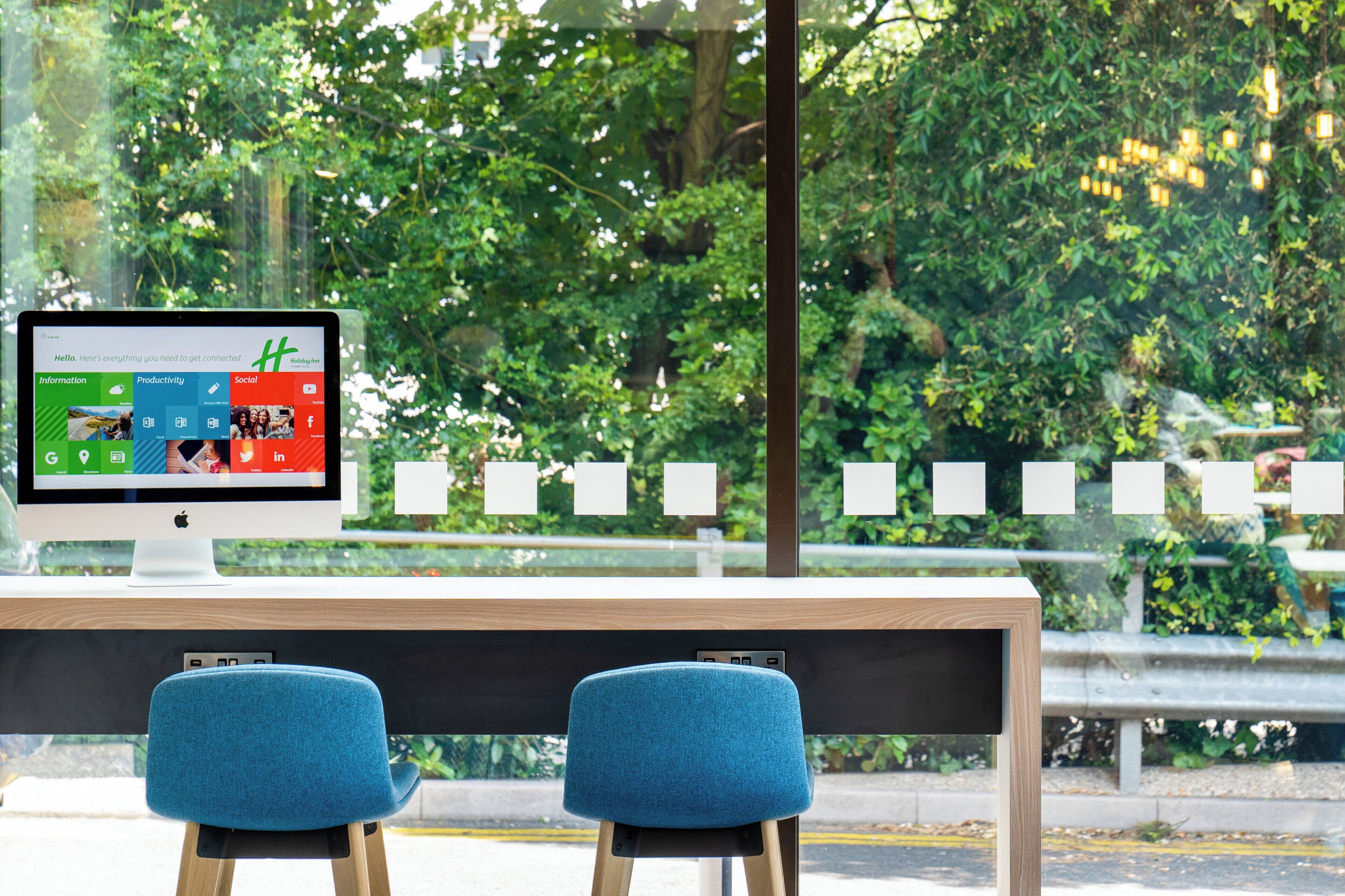 A computer monitor on a desk with a view of a park through a window.