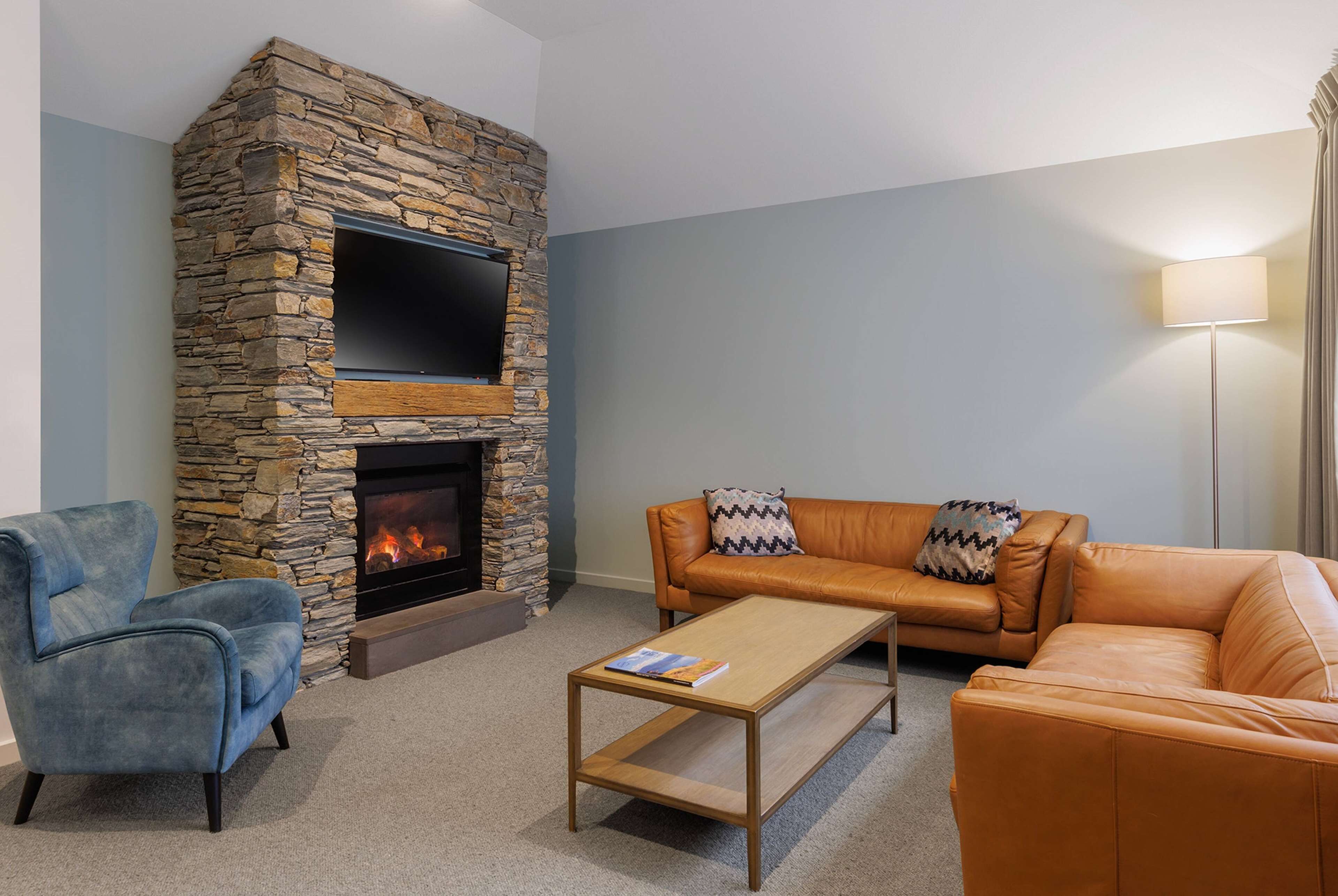 A living room with a stone fireplace, a flat-screen TV, a blue armchair, an orange leather sofa, and a coffee table.