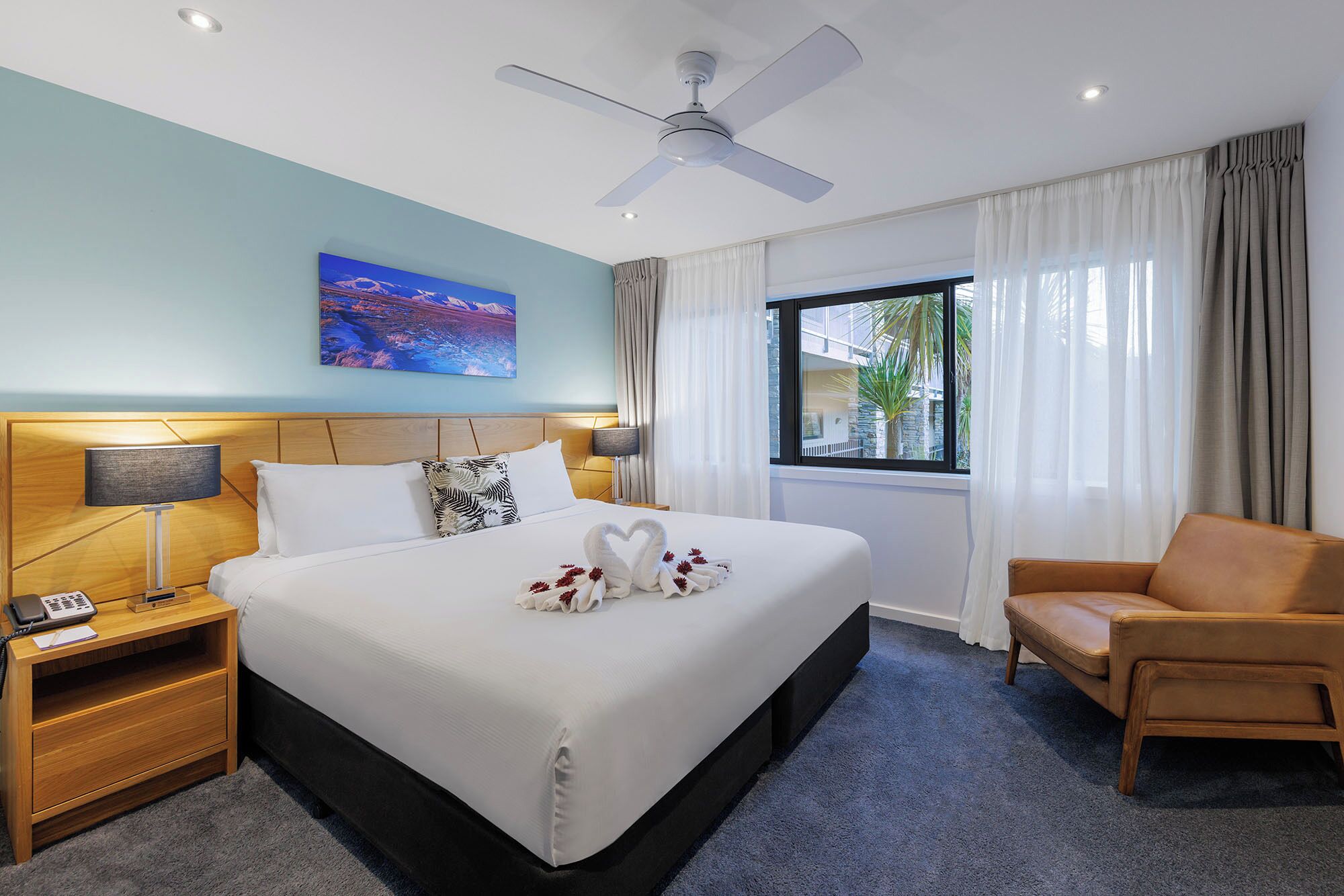 A hotel room with a bed, a nightstand, a chair, a ceiling fan, and a window with curtains.
