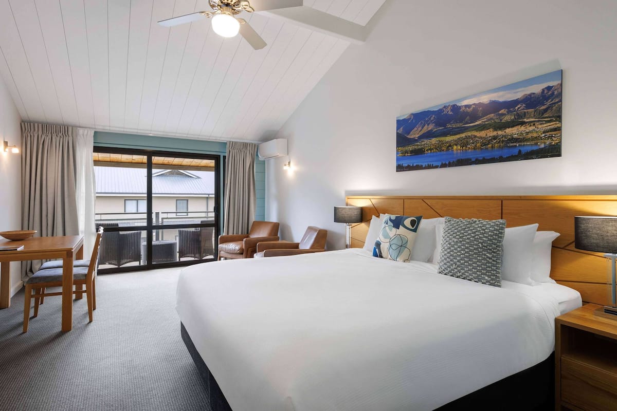 A hotel room with a large bed, a desk with chairs, a balcony with a view of mountains, and a painting on the wall.