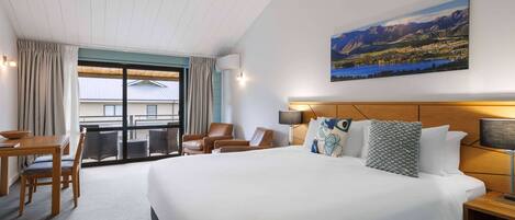 A hotel room with a large bed, a desk with chairs, a balcony with a view of mountains, and a painting on the wall.