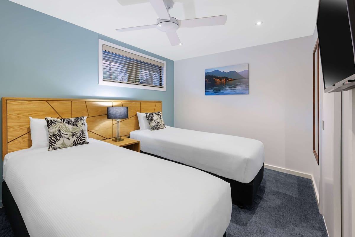 A hotel room with two beds, a ceiling fan, a window with blinds, a painting of a landscape, and a TV on the wall.
