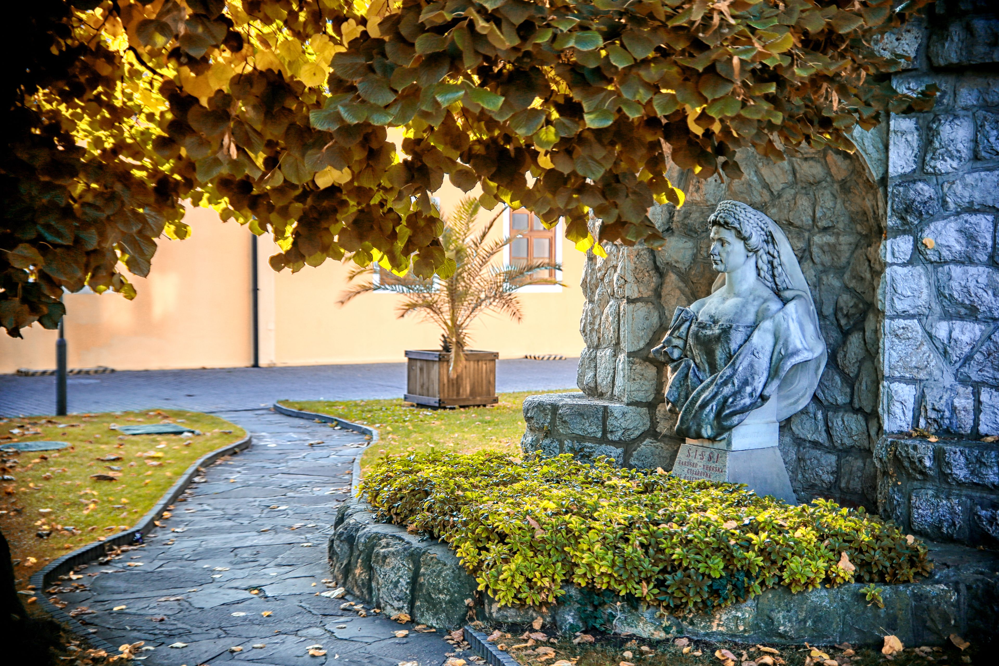 A stone pathway leading to a statue of a figure holding a book, surrounded by greenery and a stone wall.
