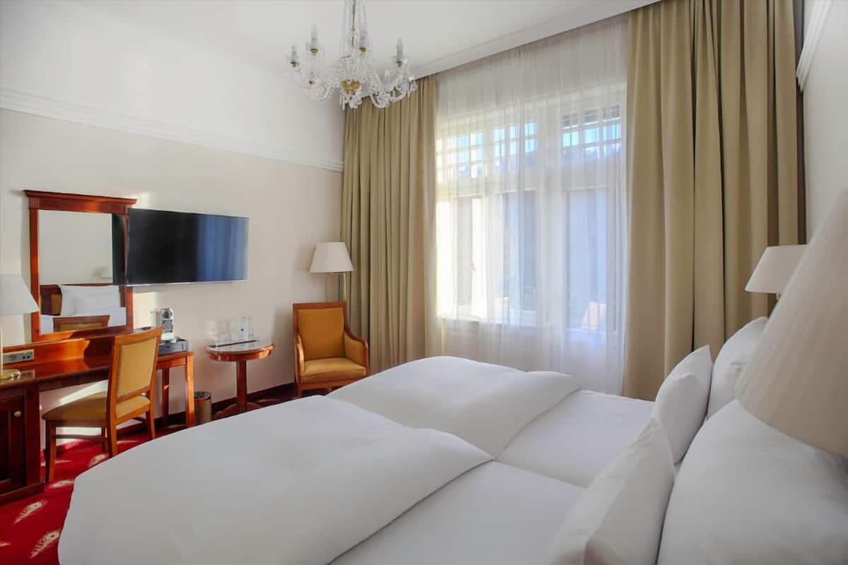A hotel room with a large bed, a desk with a flat-screen TV, a chair, and a window with curtains.