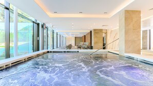 A modern indoor pool with clear glass walls, a seating area with chairs, and a marble wall. - Ensana Thermia Palace (Piestany)