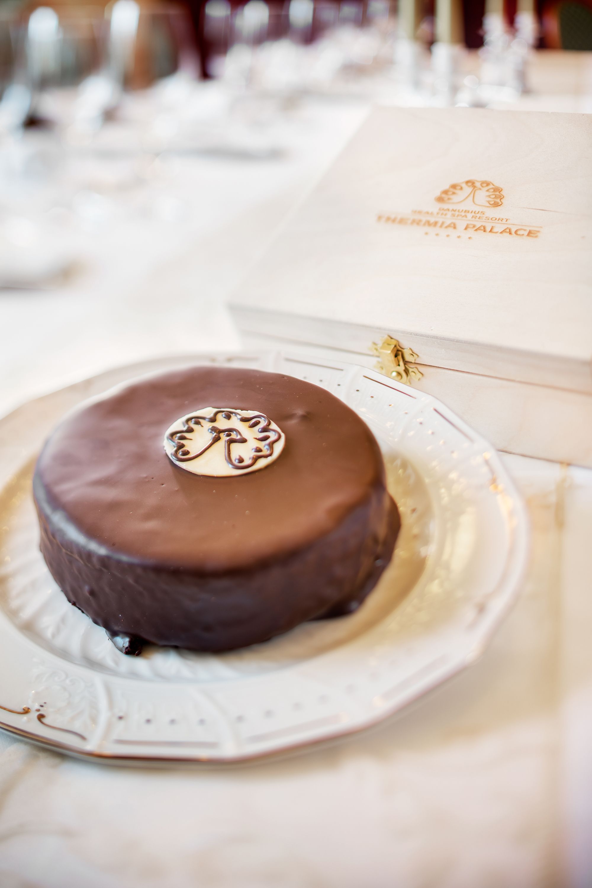 Un gâteau au chocolat sur une assiette blanche ornée d’un logo, posé sur une table recouverte d’une nappe blanche.