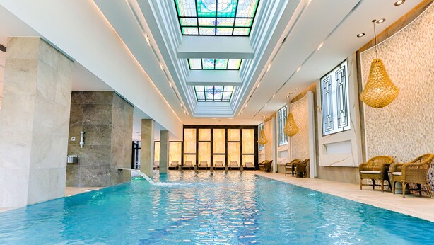 A swimming pool with a skylight ceiling, lounge chairs, and a modern interior design. - Ensana Thermia Palace (Piestany)