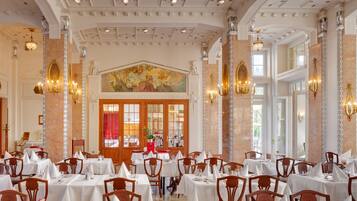 A formal dining room with multiple tables and chairs, a large mural on the wall, and classical architectural details.