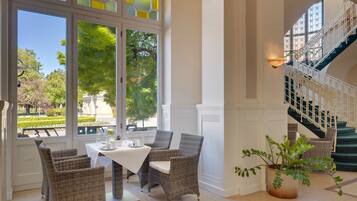 A dining area with wicker chairs and a table, large windows with a view of greenery, and a staircase with a railing.