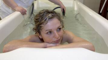 Une personne dans une baignoire remplie d’eau, une autre personne debout à côté de la baignoire, et un robinet visible.