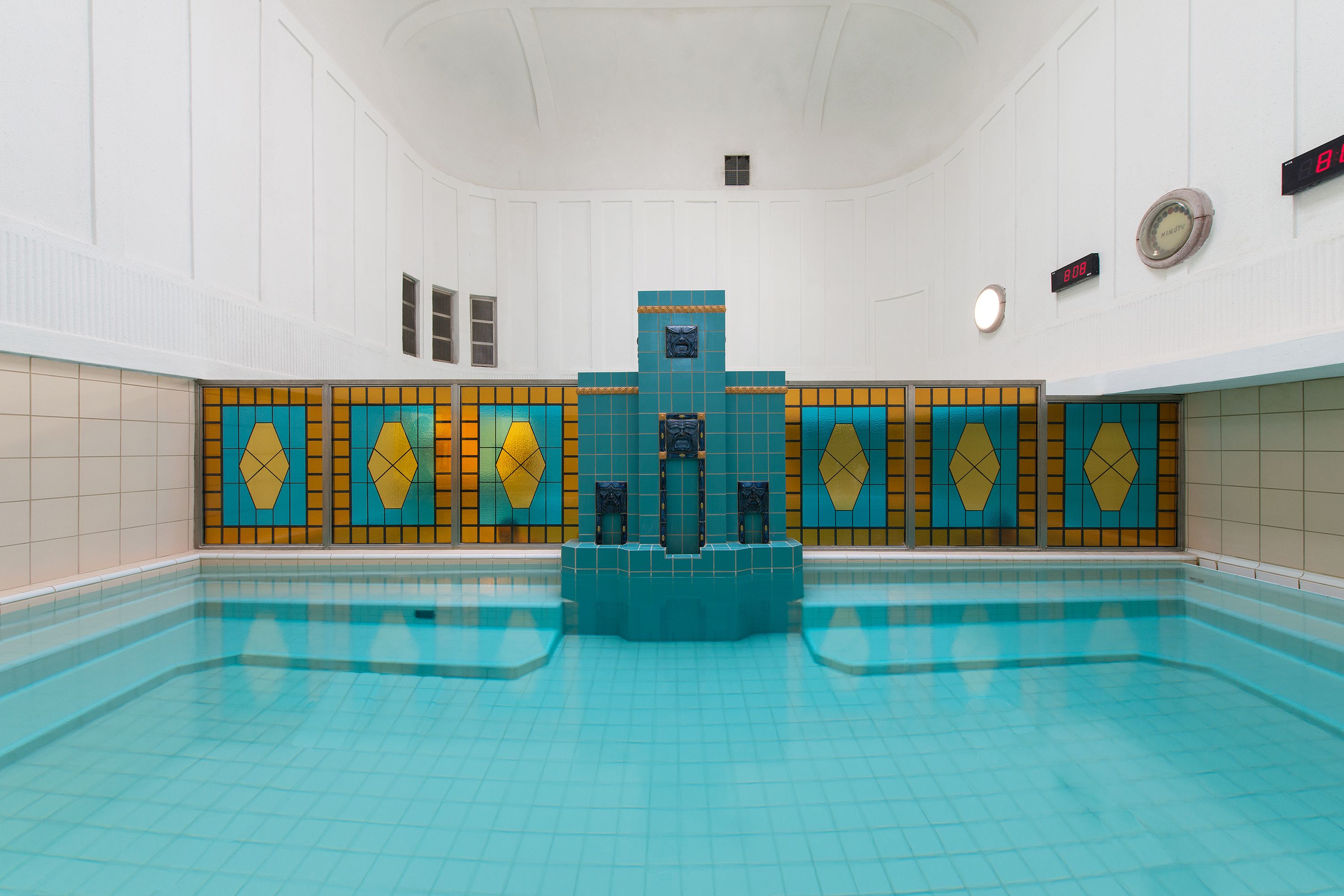 A large indoor swimming pool with a central fountain, tiled walls, and a clock on the wall.