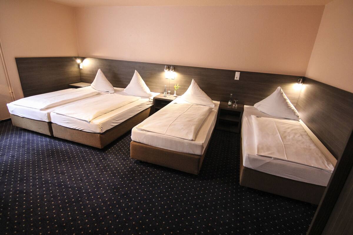 A hotel room with three single beds, each with a pillow and a small table beside it.