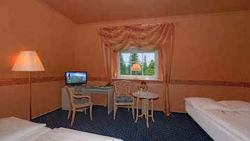 A hotel room with a bed, a desk with a computer, a chair, a small table with a vase, and a window with curtains.