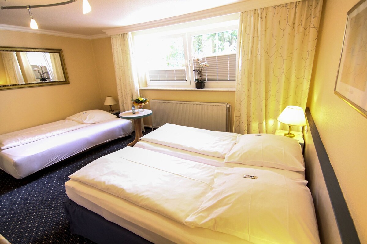 A hotel room with two beds, a small round table with a lamp, a window with curtains, and a radiator.