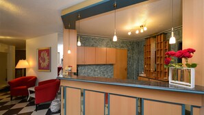 A hotel lobby with a check-in counter, red armchairs, a large mirror, and a vase with red flowers. - Hotel Nordic (Norderstedt)