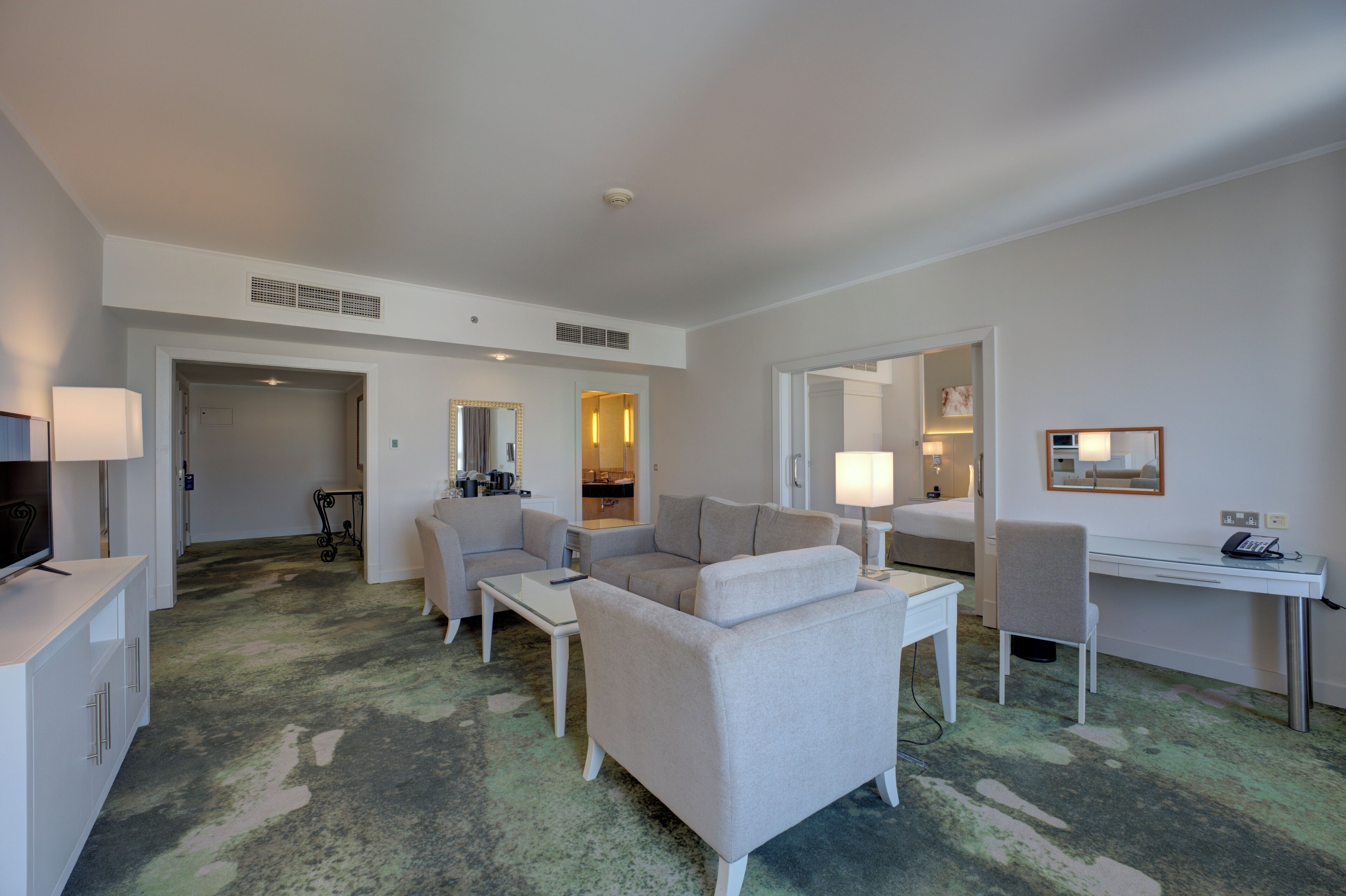 Une chambre d’hôtel dotée d’un coin salon comprenant un canapé, une table basse, un bureau et une chaise. On y trouve également un téléviseur posé sur un meuble, une lampe et un miroir au mur.
