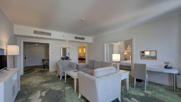 Une chambre d’hôtel dotée d’un coin salon comprenant un canapé, une table basse, un bureau et une chaise. On y trouve également un téléviseur posé sur un meuble, une lampe et un miroir au mur.
