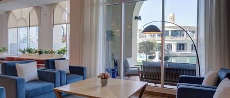 A spacious lounge area with blue sofas, a wooden coffee table, and large windows offering a view of a building with a dome and a pool.